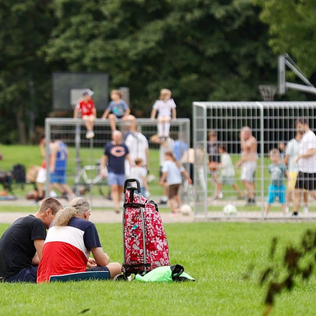 Viele Kinder und Jugendliche sitzen auf einer Wiese, im Hintergrund spielen einige Fußball auf einem Bolzplatz.