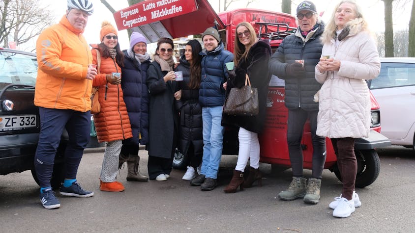 Eine Gruppe Menschen stehen im Halbkreis vor einem feuerroten Kaffeemobil.