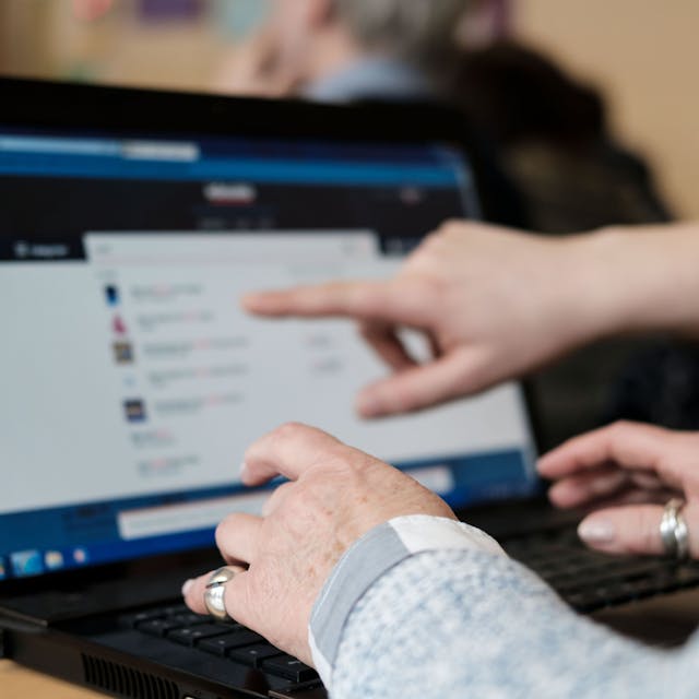 Eine Teilnehmerin des Computerkurses für Senioren übt zusammen mit einem Schüler an einem Laptop. Sie hält ihre Hände über die Tastatur, während ein Schüler mit einem Finger auf den Bildschirm zeigt.