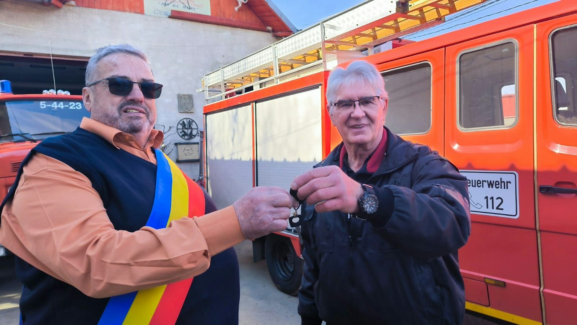 Winfried Dederichs gibt Sorin Ionita Vasile einen Schlüssel. Die beiden stehen vor einem Feuerwehrauto, Vasile trägt eine Schärpe in den Farben der rumänischen Flagge.