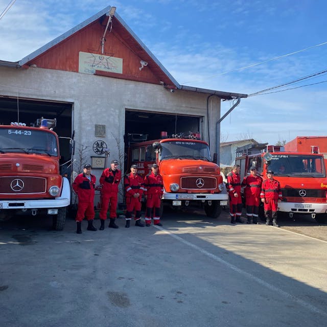 Rot gekleidete Feuerwehrleute stehen vor drei roten Feuerwehrwagen.