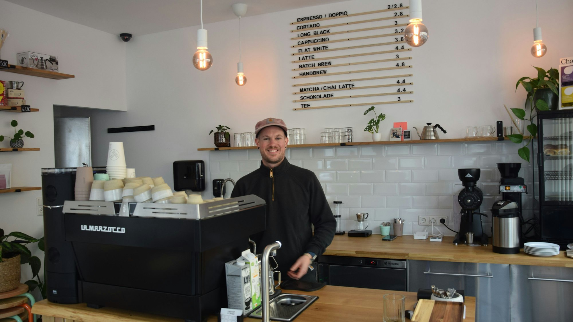 Ein junger Mann mit Kappe steht hinter eine Kaffeemaschine in seinem Café.