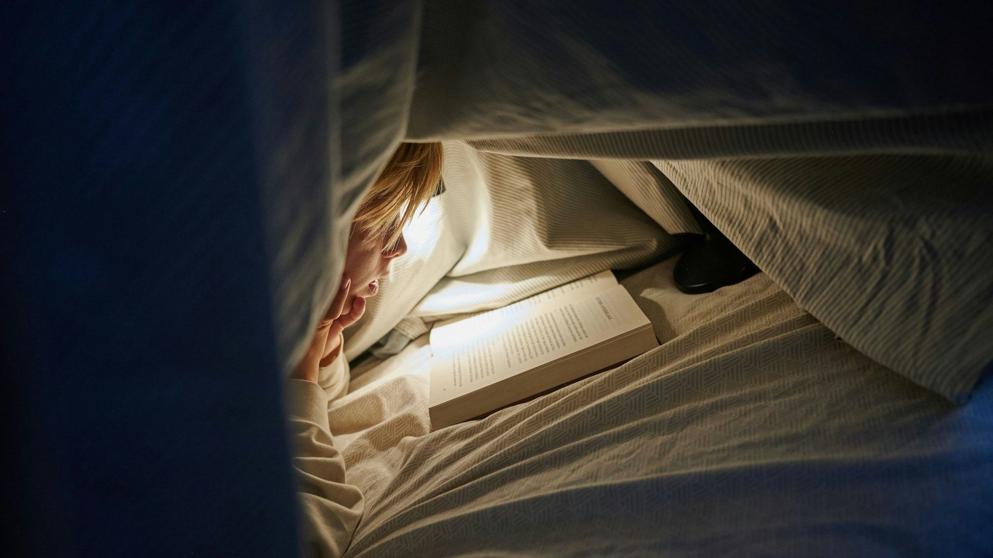 Ein Kind liegt auf einem Bett in einem dunklen Zimmer und liest unter einer Bettdecke mit einer Lampe einBuch.