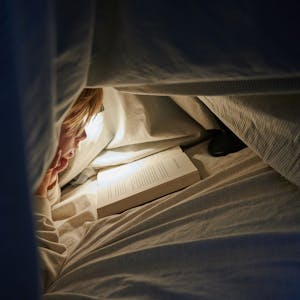 Ein Kind liegt auf einem Bett in einem dunklen Zimmer und liest unter einer Bettdecke mit einer Lampe ein&nbsp;Buch.&nbsp;