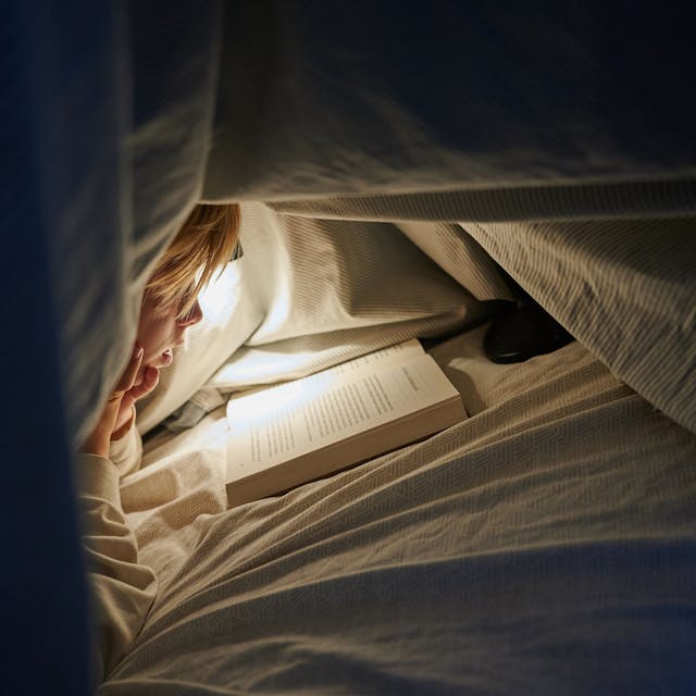 Ein Kind liegt auf einem Bett in einem dunklen Zimmer und liest unter einer Bettdecke mit einer Lampe ein&nbsp;Buch.&nbsp;