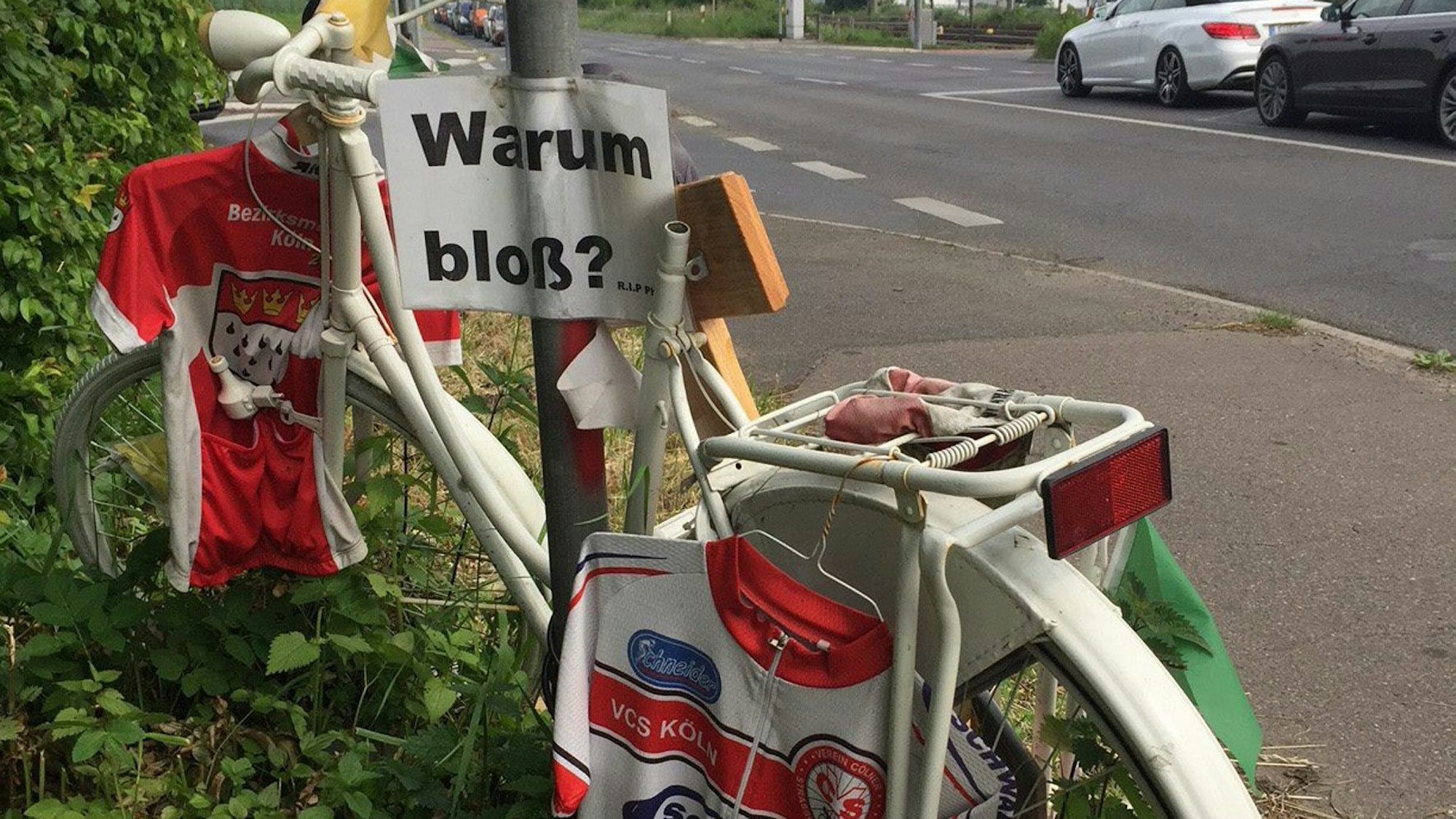 Trikots hängen an einem weißen Fahrrad. Am Schildermast hängt ein Schild mit der Aufschrift „Warum bloß?“.