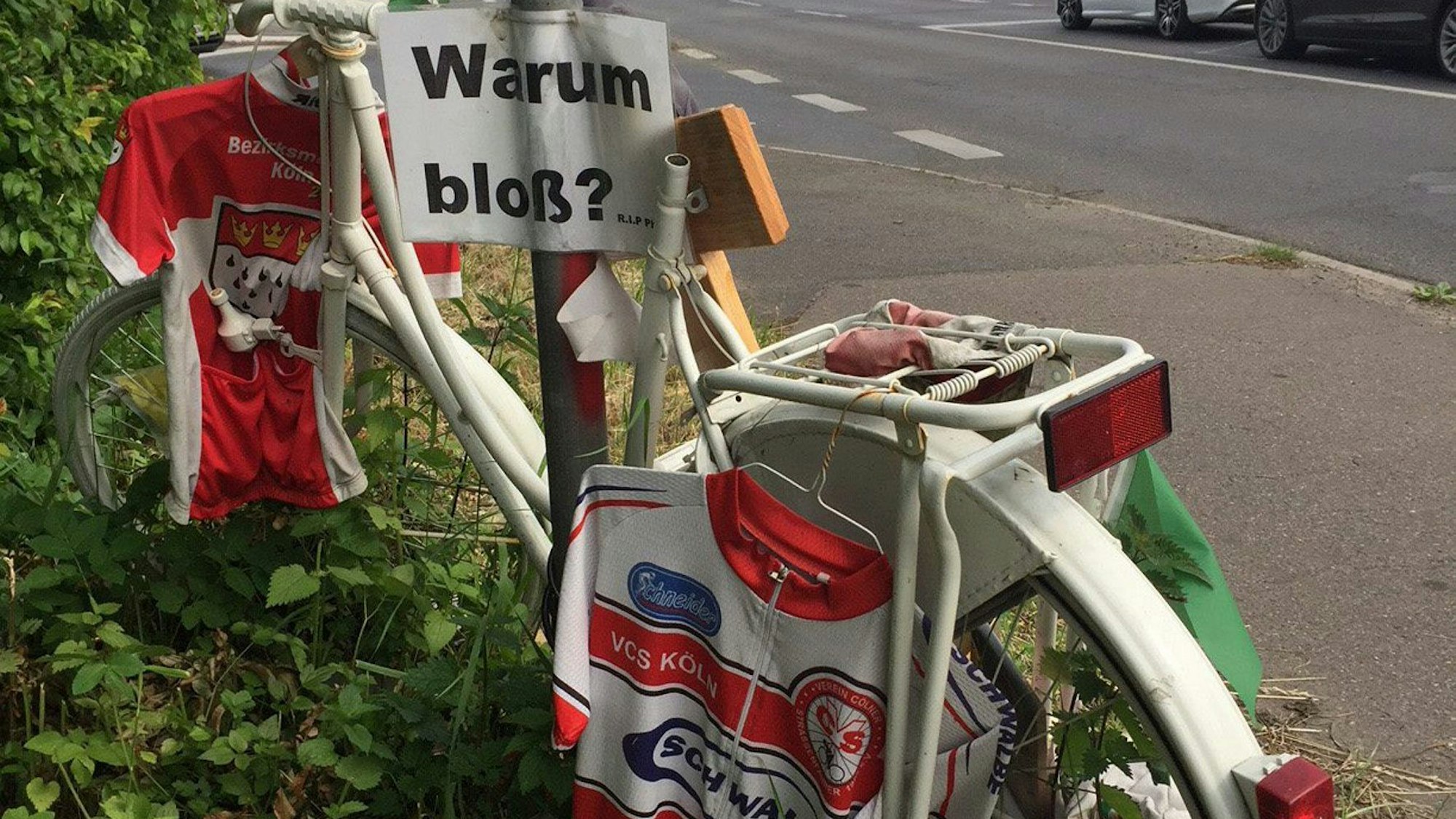 Trikots und ein Schild mit der Aufschrift „Warum bloß?“ hängen an einem weißen Fahrrad, das als Mahnmal am Straßenrand aufgestellt ist.