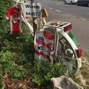 Trikots hängen an einem weißen Fahrrad. Am Schildermast hängt ein Schild mit der Aufschrift „Warum bloß?“.