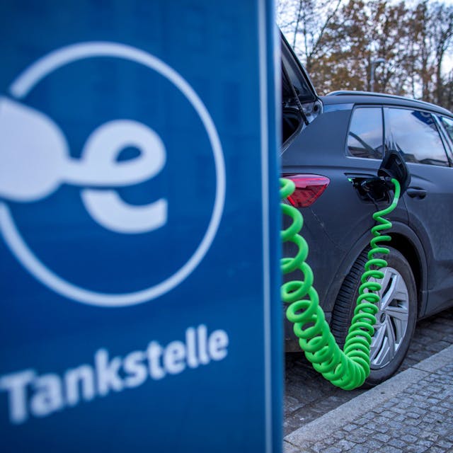 Das Symbolbild zeigt ein Elektrofahrzeug, das an einer blauen Ladesäule mit der Aufschrift „Tankstelle“ steht.