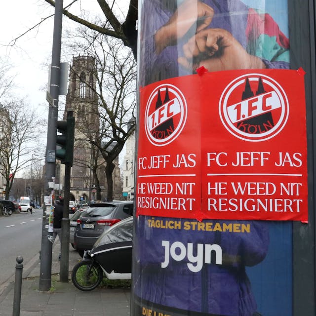 Auf einer Litfaßsäule kleben rote Plakate des 1. FC Köln mit der Aufschrift: „FC Jeff Jas – He weed nit resigniert“.&nbsp;