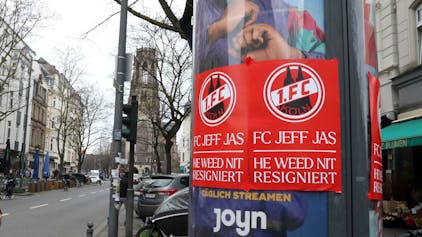 Auf einer Litfaßsäule kleben rote Plakate des 1. FC Köln mit der Aufschrift: „FC Jeff Jas – He weed nit resigniert“.&nbsp;