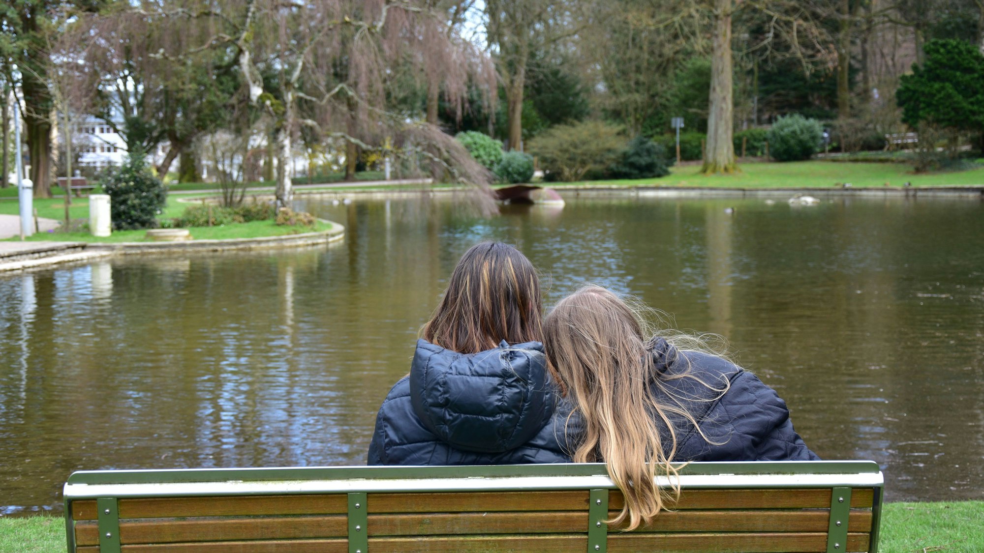 Zwei Frauen sitzen auf einer Parkbank an einem Teich, mit dem Rücken zur Kamera gewandt.