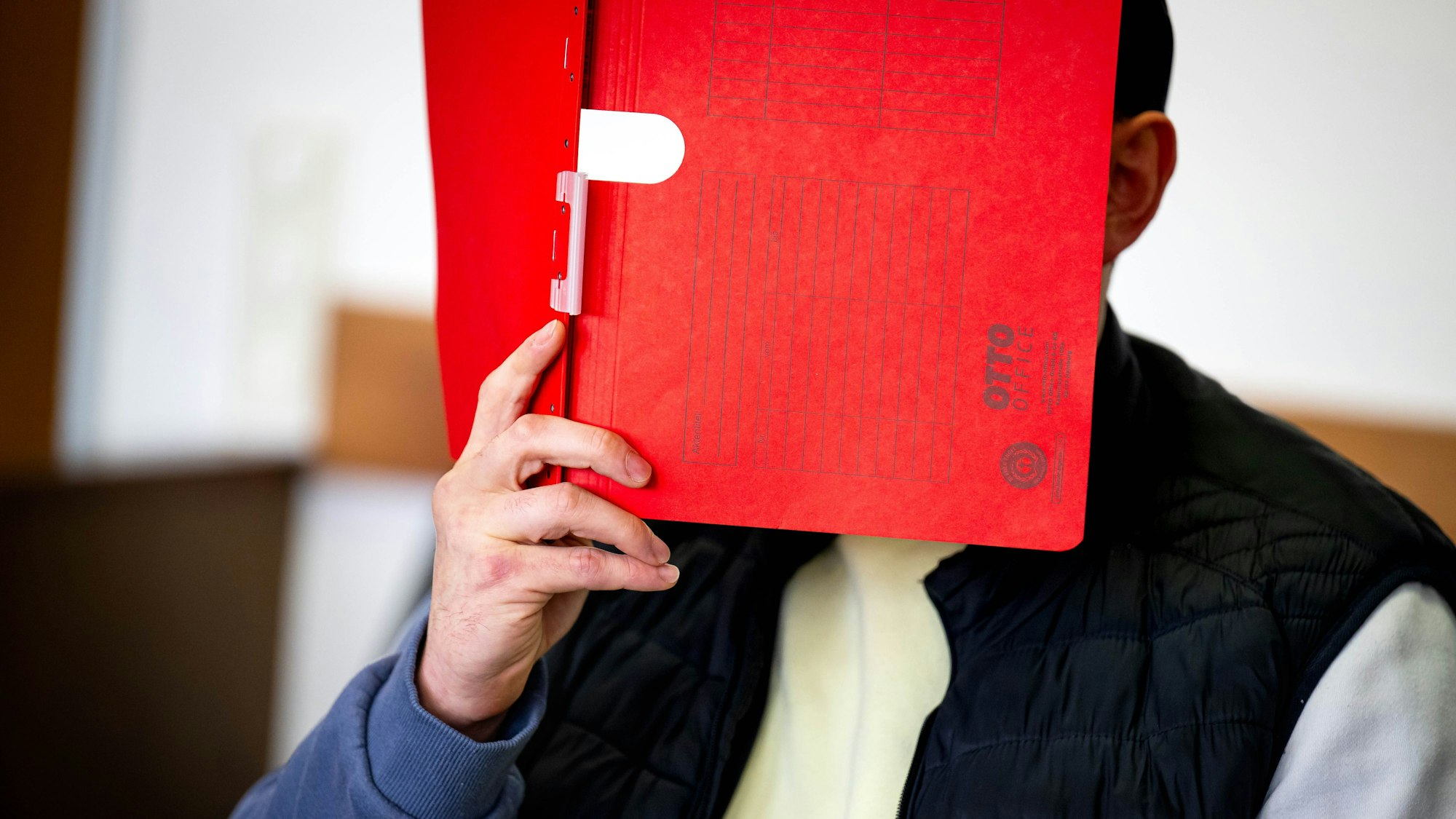 Ein Angeklagter hält sich im Gerichtssaal eine rote Mappe vor das Gesicht