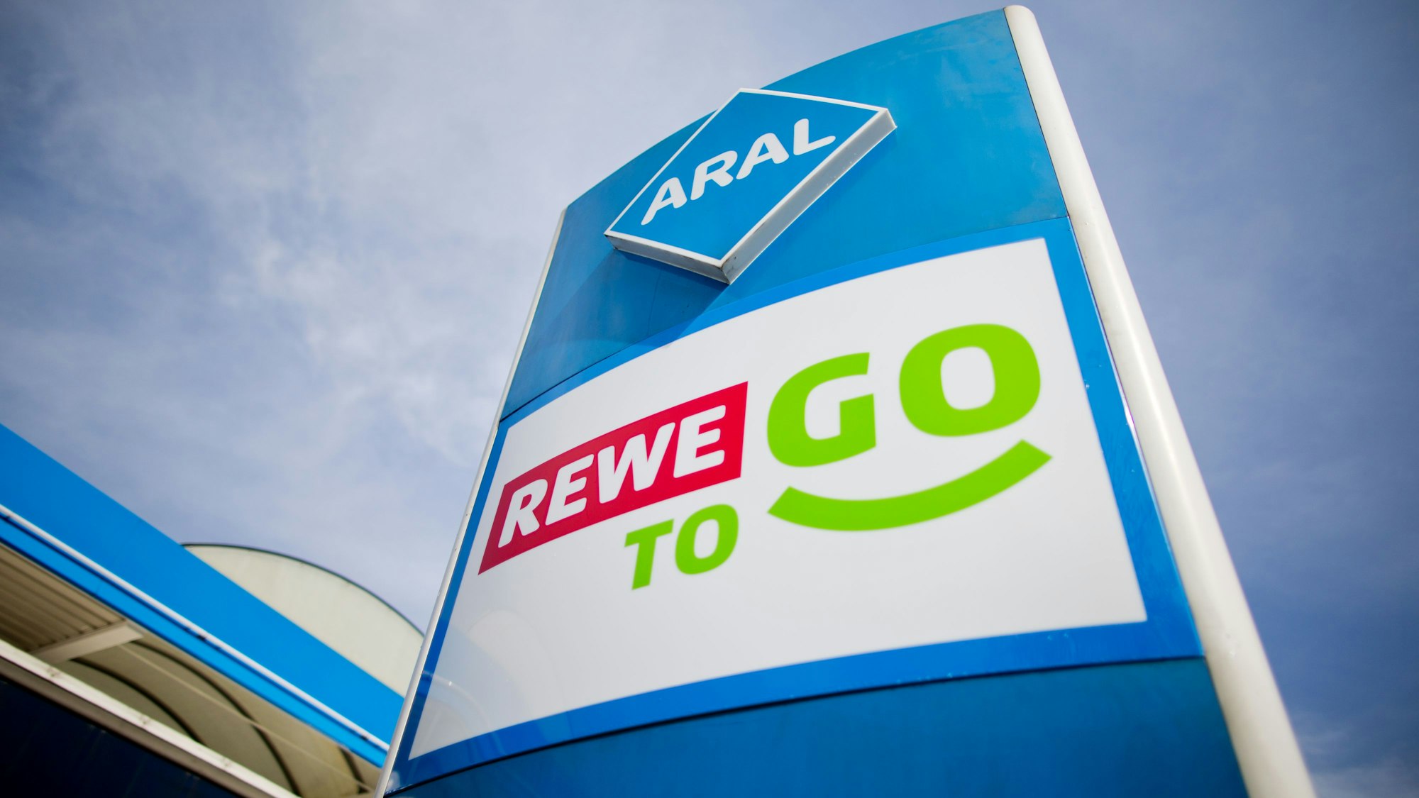 Die Logos von Rewe To Go und Aral stehen übereinander vor einer Tankstelle.