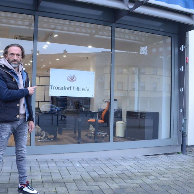 Ein Mann steht vor einem leeren Ladenlokal. Er zeigt auf ein Schild am Fenster mit der Aufschrift „Troisdorf hilft“.