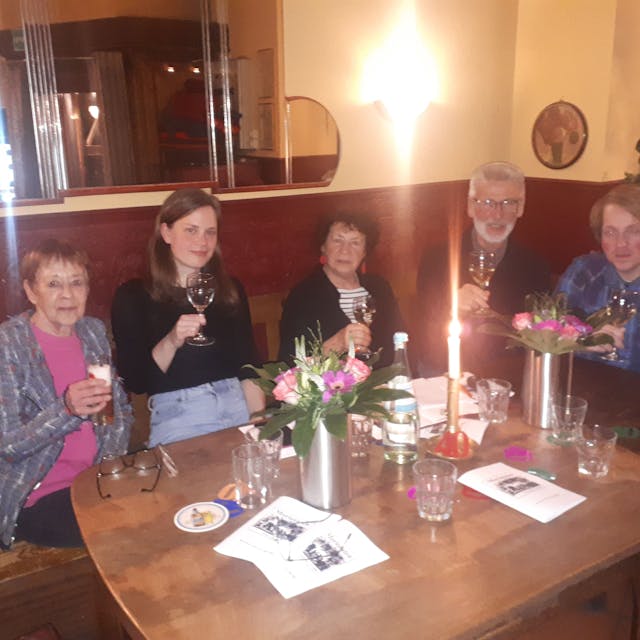 Fünf Personen sitzen mit erhobenen Gläsern an einem Tisch in einer Weinstube.