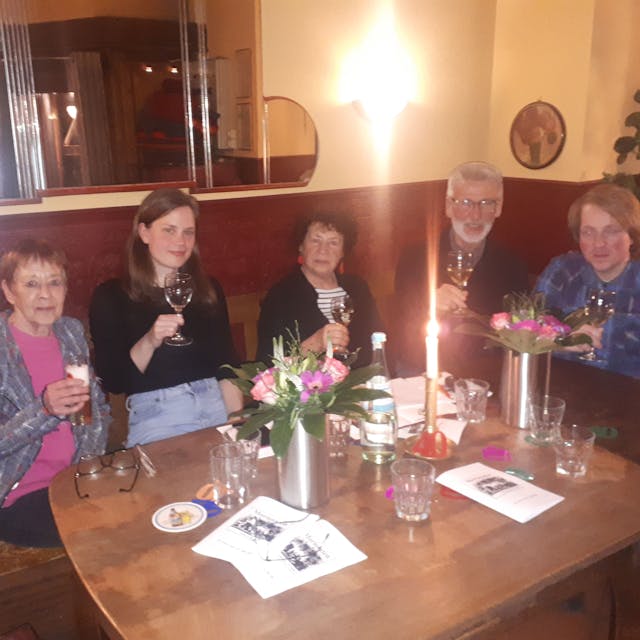 Fünf Personen sitzen mit erhobenen Gläsern an einem Tisch in einer Weinstube.