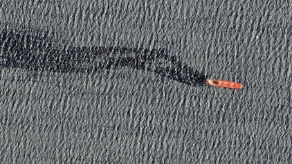 Bild von oben: Ein rotes Schiff fährt auf dem Meer. Der Frachter hinterlässt einen großen schwarzen Ölteppich hinter sich.
