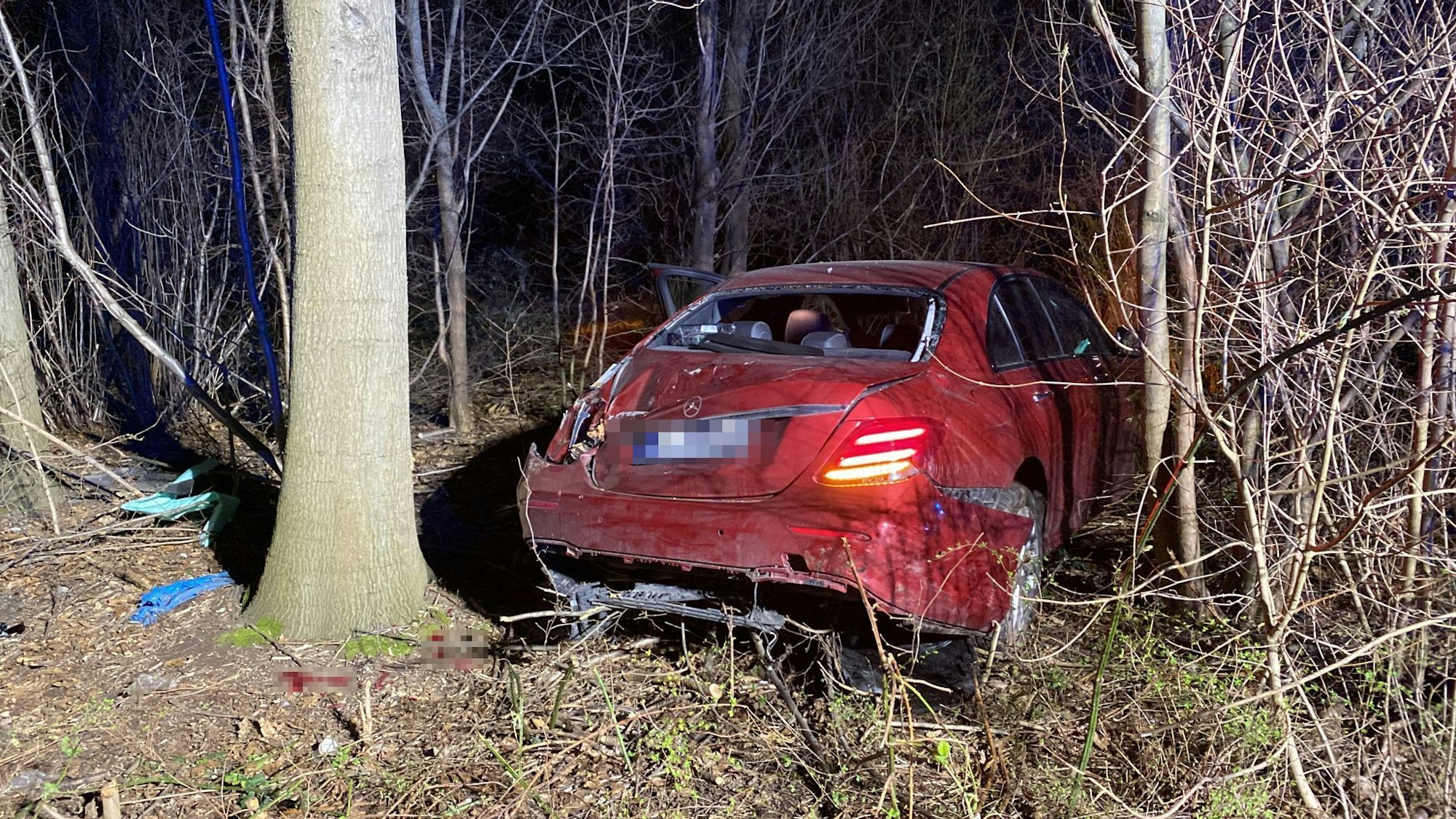 Ein rotes Auto steht zwischen Bäumen und Sträuchern. Der Wagen hat erhebliche Schäden und kaputte Scheiben.