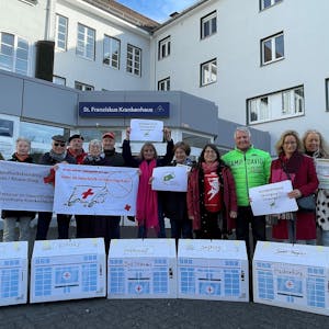 Menschen stehen vor dem Eingang des St. Franziskus-Krankenhaus Eitorf, vor ihnen Häuschen mit den Ortsnamen der bereits geschlossenen Geburtsstationen in der Region.