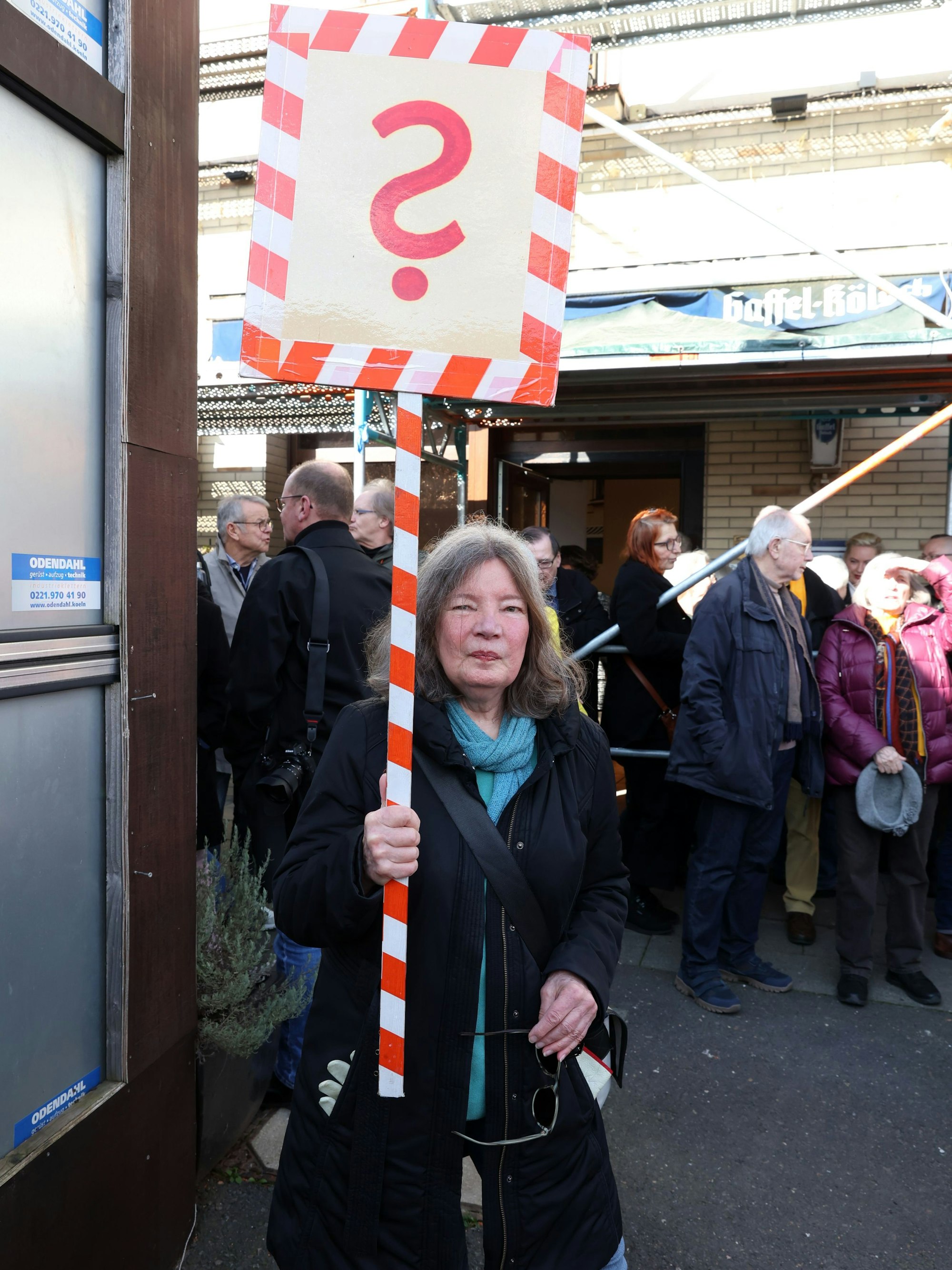 Eine Frau hält ein Schild mit einem roten Fragezeichen in der Hand und blickt in die Kamera.