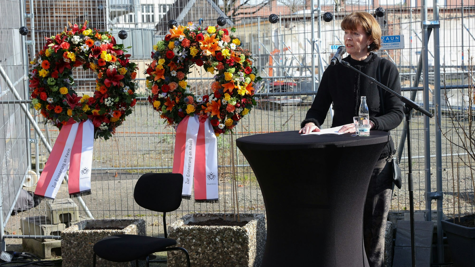 Oberbürgermeisterin Henriette Reker steht an einem Tisch und spricht in ein Mikrofon. Hinter ihr ist ein Bauzaun, an dem zwei Blumenkränze befestigt sind.