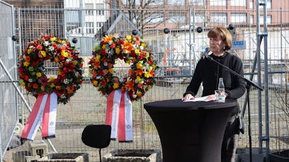 Oberbürgermeisterin Henriette Reker steht an einem Tisch und spricht in ein Mikrofon. Hinter ihr ist ein Bauzaun, an dem zwei Blumenkränze befestigt sind.&nbsp;