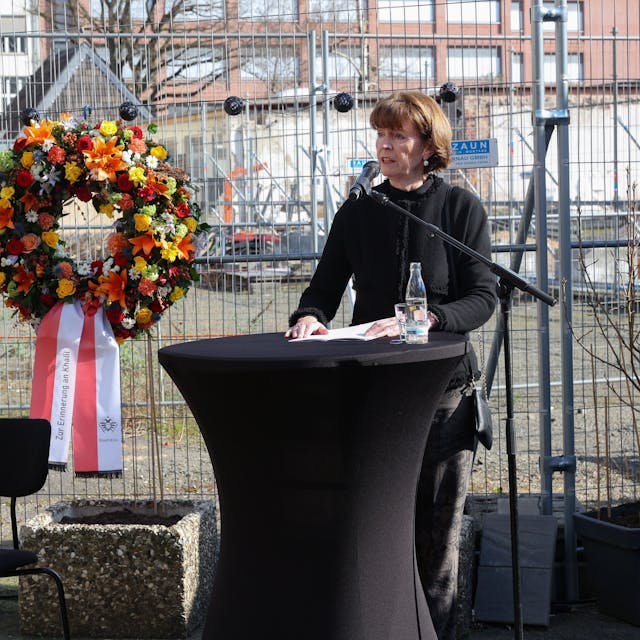Oberbürgermeisterin Henriette Reker steht an einem Tisch und spricht in ein Mikrofon. Hinter ihr ist ein Bauzaun, an dem zwei Blumenkränze befestigt sind.&nbsp;