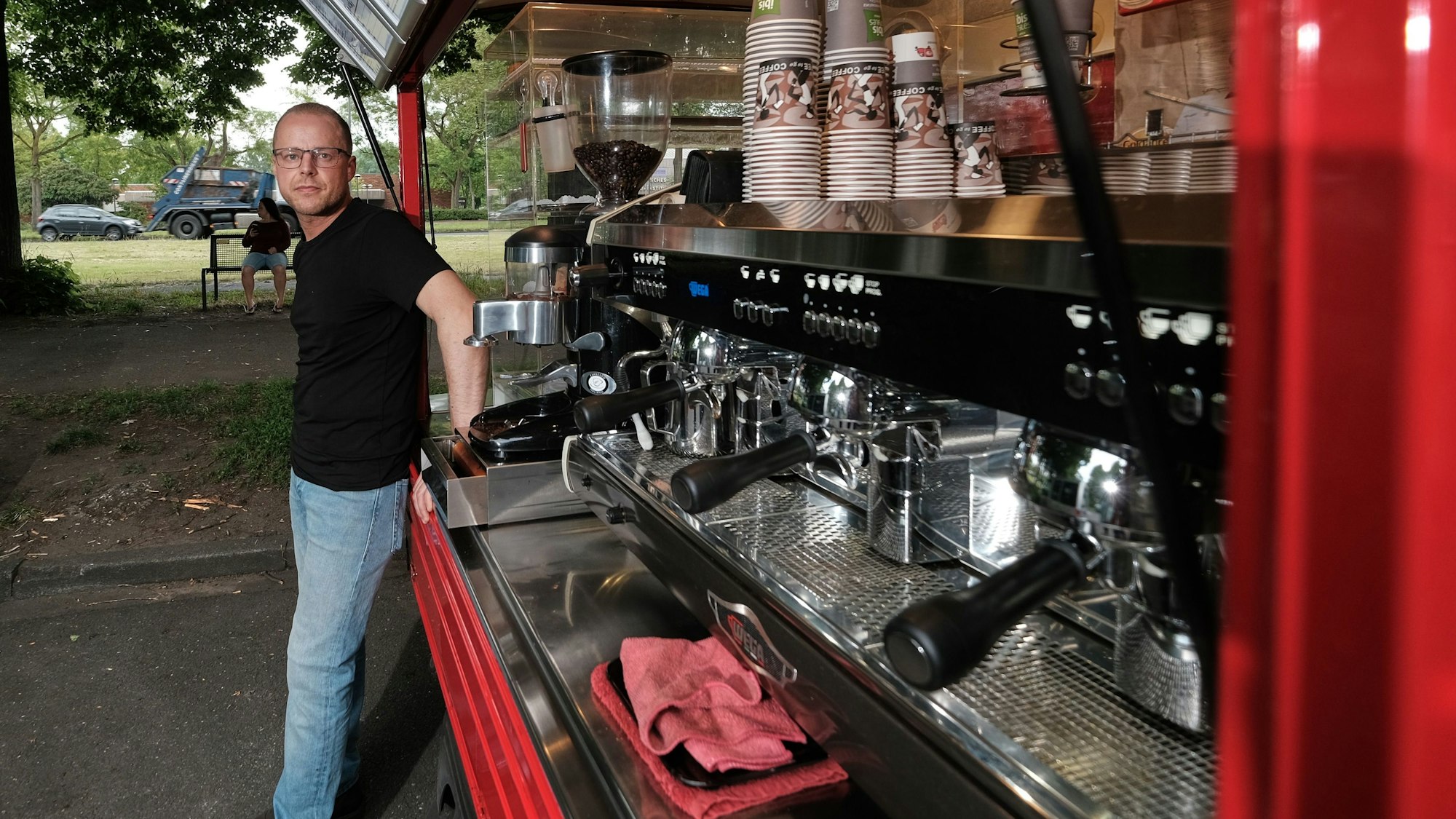 Roberto Mirabile steht vor seinem geöffneten Caféccino-Mobil, in dem er Kaffeespezialitäten und Gebäck anbietet. Zu sehen sind drei Siebträgermaschinen, die nebeneinander angebracht sind, im Hintergrund sind Behälter mit Kaffeebohnen sichtbar. Oberhalb der Kaffeemaschinen sind Pappbecher aufgereiht.