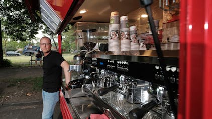 Roberto Mirabile steht vor seinem geöffneten Caféccino-Mobil, in dem er Kaffeespezialitäten und Gebäck anbietet. Zu sehen sind drei Siebträgermaschinen, die nebeneinander angebracht sind, im Hintergrund sind Behälter mit Kaffeebohnen sichtbar. Oberhalb der Kaffeemaschinen sind Pappbecher aufgereiht.&nbsp;