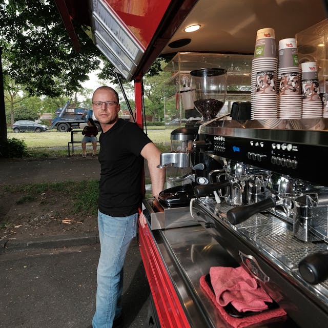 Roberto Mirabile steht vor seinem geöffneten Caféccino-Mobil, in dem er Kaffeespezialitäten und Gebäck anbietet. Zu sehen sind drei Siebträgermaschinen, die nebeneinander angebracht sind, im Hintergrund sind Behälter mit Kaffeebohnen sichtbar. Oberhalb der Kaffeemaschinen sind Pappbecher aufgereiht.&nbsp;