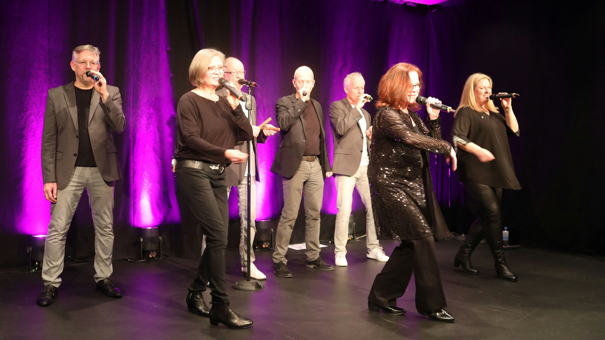 Eine Acapella-Musikgruppe bestehend aus sieben Sängern und Sängerinnen steht auf der Bühne. Jede Person hält ein Mikrofon in der Hand.