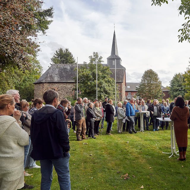 Zahlreiche Menschen stehen bei einer Bürgerversammlung in Heimbach-Blens auf einer Wiese, im Hintergrund ist die Kirche zu sehen.