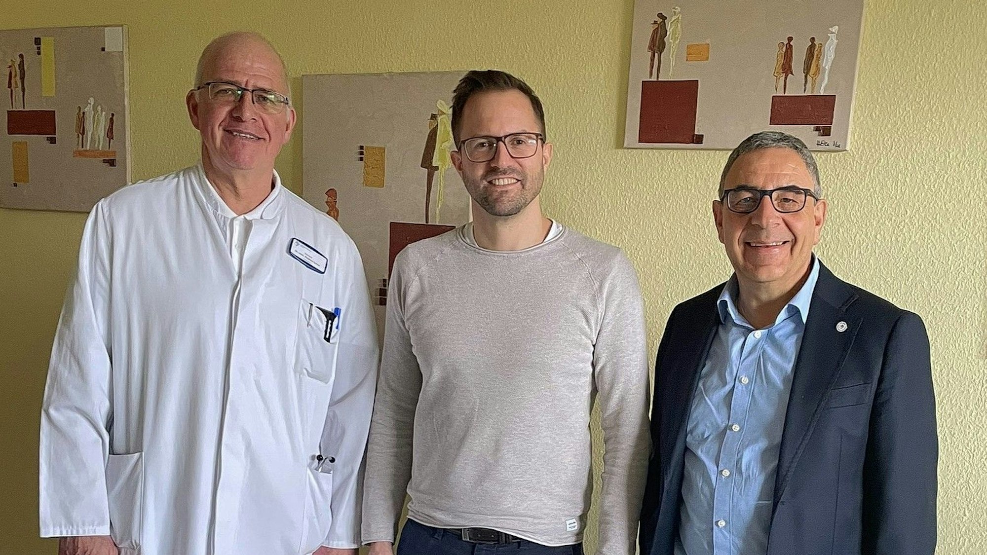 Drei Männer mit Brille stehen vor einer Wand, einer trägt einen Arztkittel.