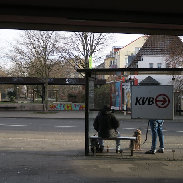 An einer Bushaltestelle warten zwei Fahrgäste, einer hat einen Hund dabei.