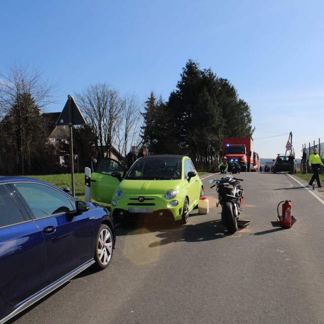 Beschädigte Zweiräder und ein Auto stehen auf einer Fahrbahn, auf dem Boden ist Ölbindemittel zu sehen. Im Hintergrund stehen Einsatzkräfte der Feuerwehr.