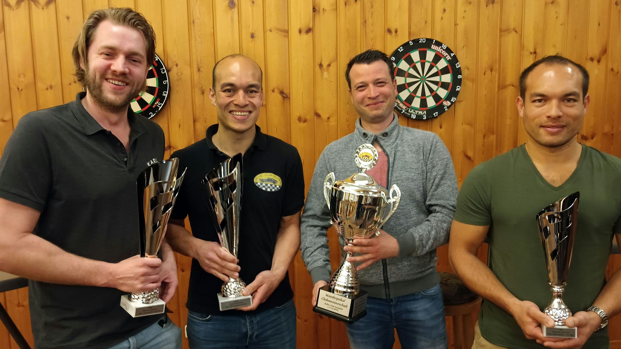 Fabian Schumacher (v.l.), Michael Klein, Andreas Nocker und Daniel Klein stehen vor einer Holzwand mit einem Dartboard und halten verschiedene Siegerpokale in den Händen.