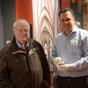 Rolf Müller-Blagovich und Sacha Reichelt stehen im Stadtmuseum Euskirchen. Reichelt zeigt eine der Münzen, die Müller der Stadt geschenkt hat.