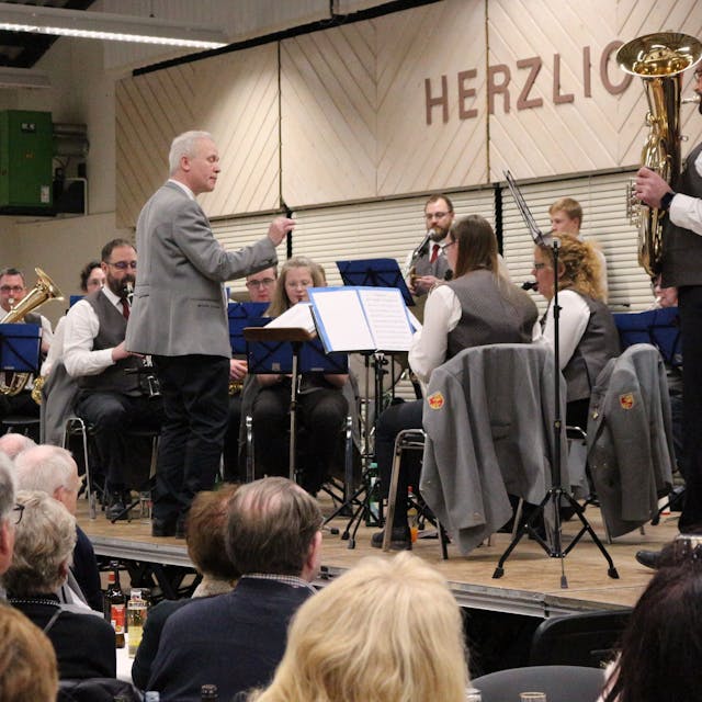 Das Orchester spielt auf der Bühne der Schwerfener Schützenhalle, dirigiert von Rudi Wedel. Im Vordergrund sieht man Teile des Publikums.