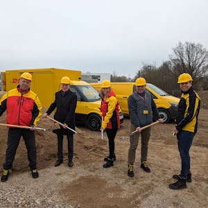 Vier Männer und eine Frau mit gelben Helmen und Spaten auf der Baustelle. Im Hintergrund gelbe Zustellfahrzeuge der Post.