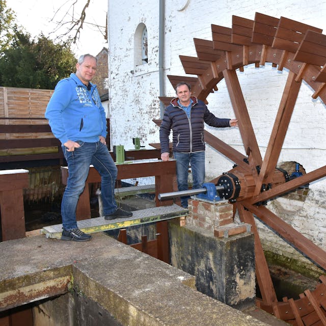 Zwei Männer stehen an einem hölzernen Mühlrad einer Wassermühle.