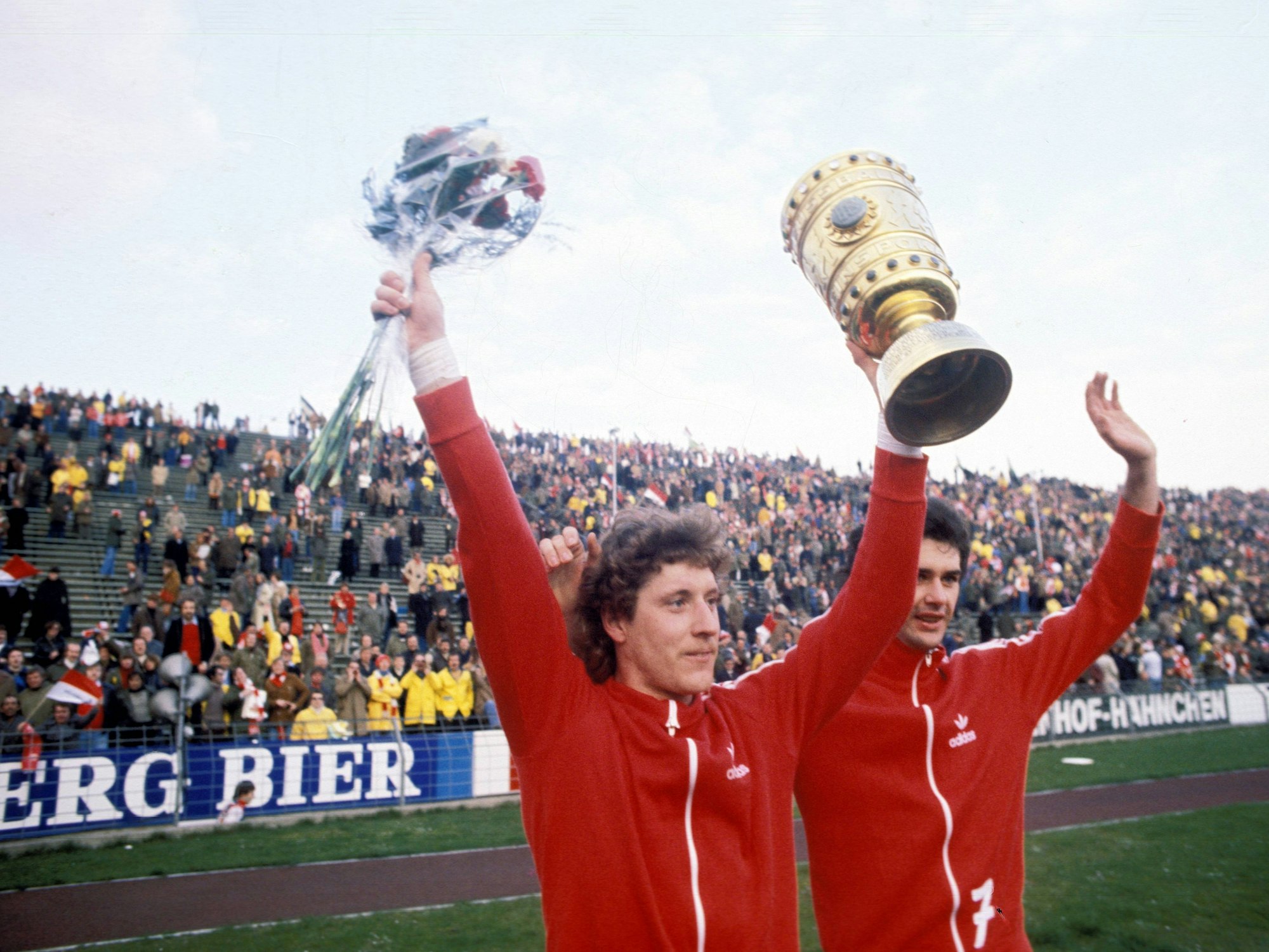 Toni Schumacher mit beiden Händen nach oben gestreckt, in der linken Hand den DFB-Pokal in der rechten einen Strauß Blumen. Rechts von ihm sein Teamkollege Gerd Strack.