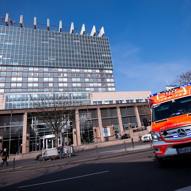 Zu sehen sind das Bettenhaus der Kölner Uniklinik und ein Rettungswagen in der Vorderansicht.