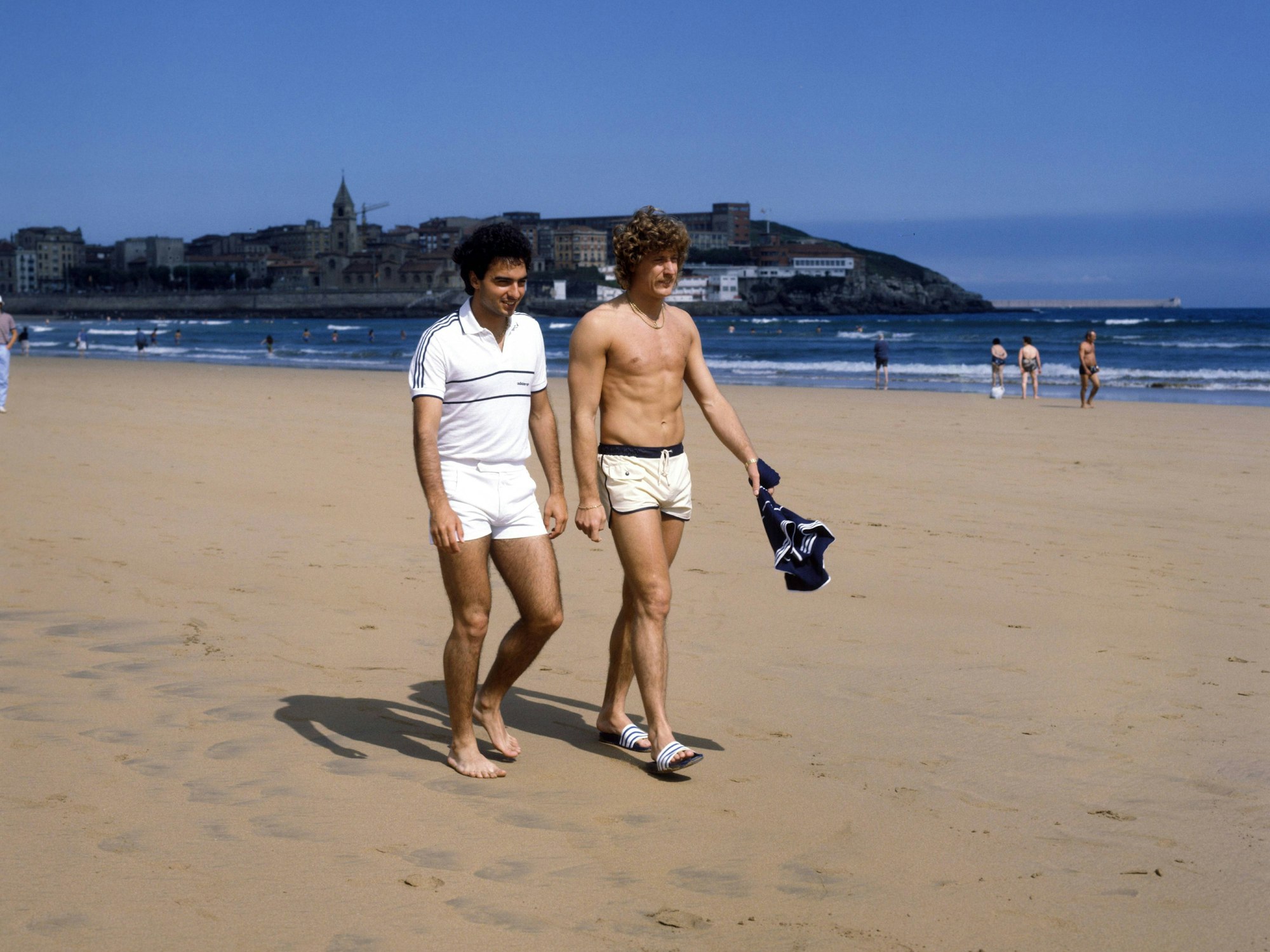 Toni Schumacher (oberkörperfrei) und Hans Müller (mit weißem Poloshirt) machen einen Spaziergang am Strand.
