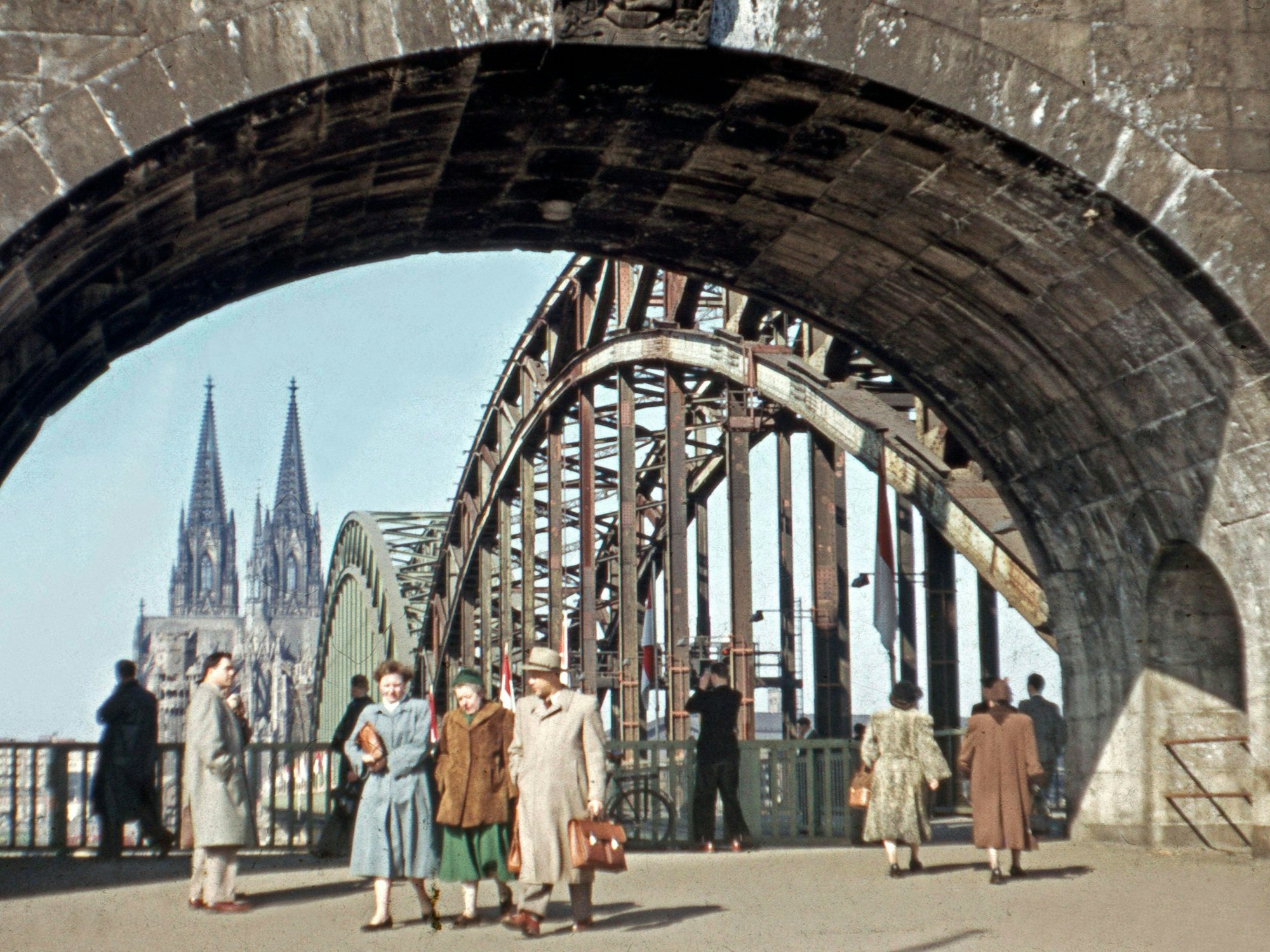 Passanten und Passantinnen flanieren unter dem Brückenportal der Hohenzollernbrücke in Köln. Im Hintergrund ist der Kölner Dom zu sehen.