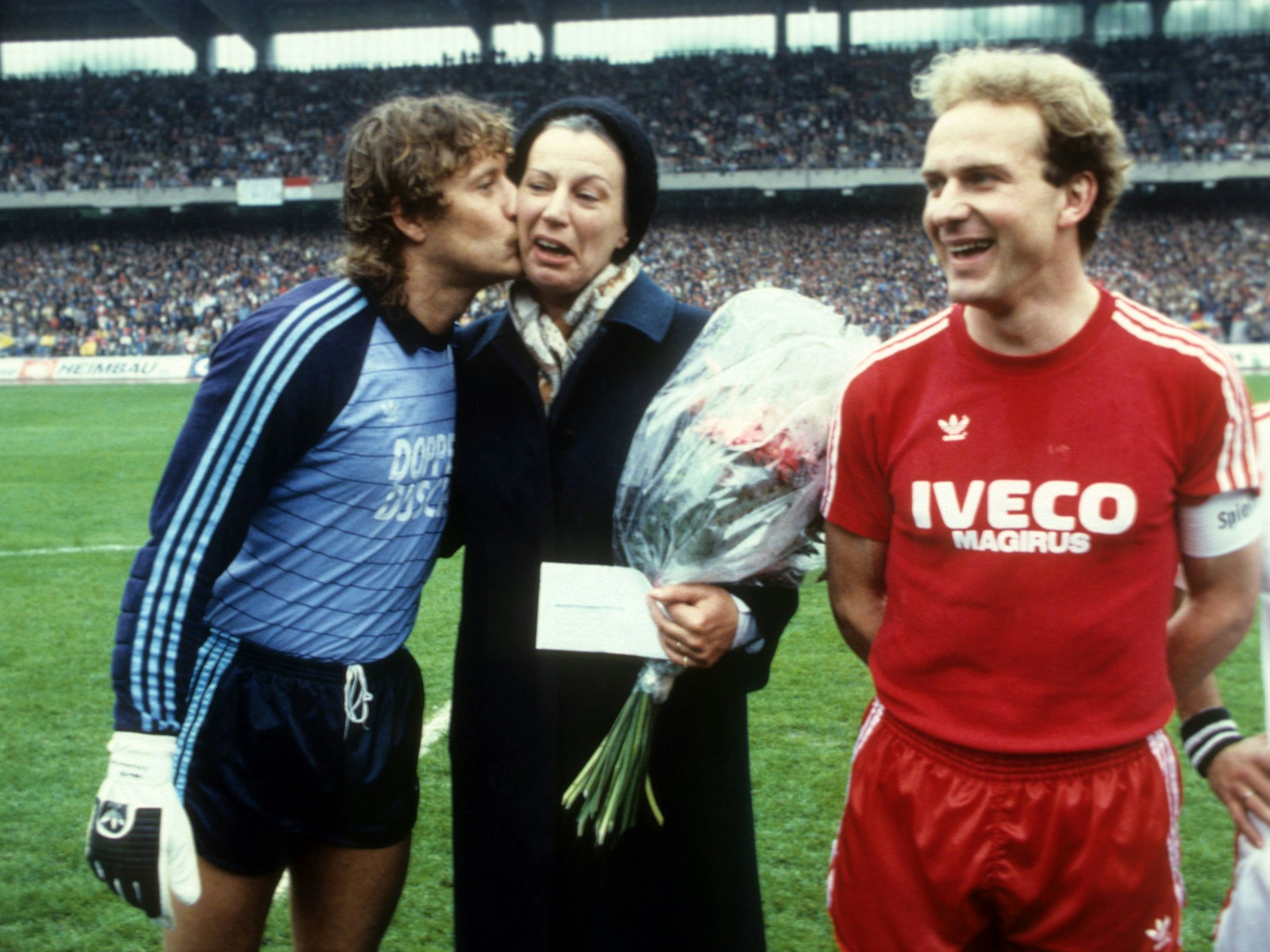 Toni Schumacher gibt Mildrer Scheel einen Kuss auf die Wange. Rechts daneben lacht Karl-Heinz Rummenigge vom FC Bayern München.