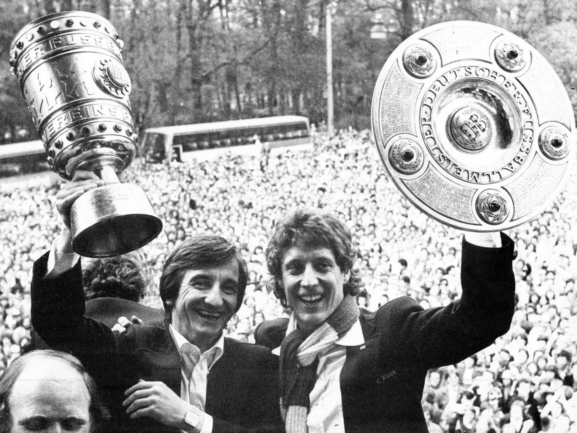 Toni Schumacher hält auf dem Rathausbalkon in Köln die Meisterschale in die Höhe, links von ihm Hannes Lähr den Pokal.