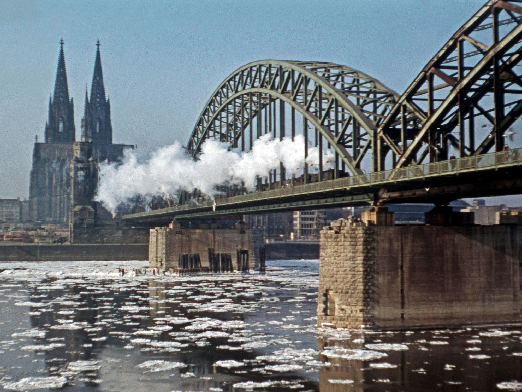 Eisschollen treiben unter der Hohenzollernbrücke auf dem Rhein, zudem überquert eine Dampflok die Brücke.