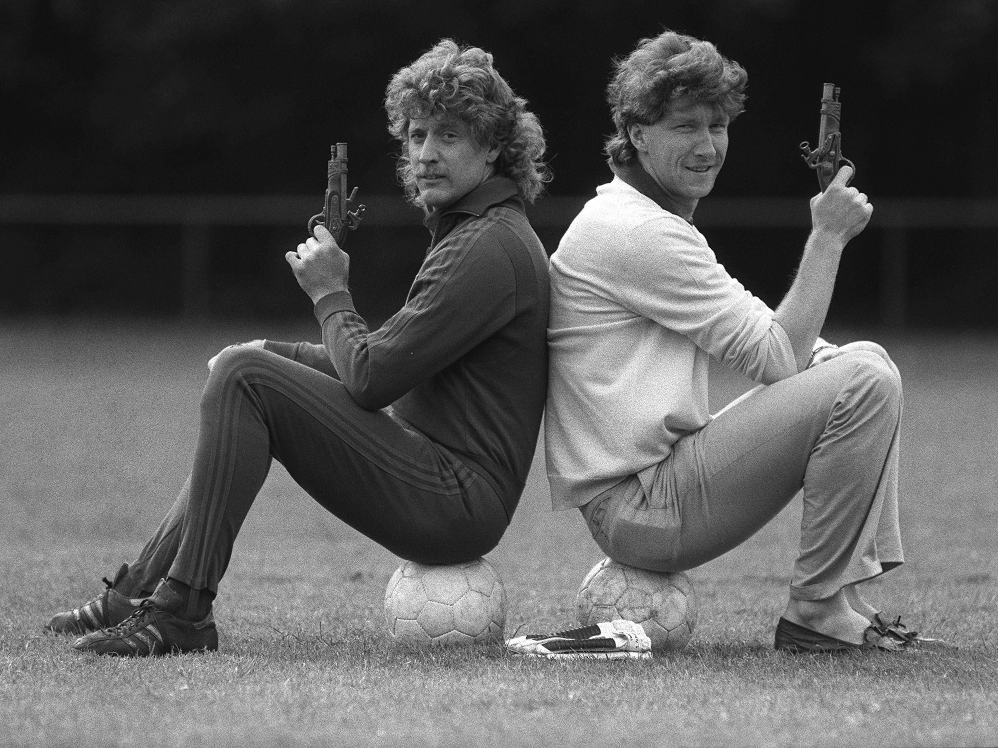 Toni Schumacher und Dieter Schatzschneider sitzen Rücken an Rücken jeder auf einem Ball, in der Hand jeweils eine Pistole.