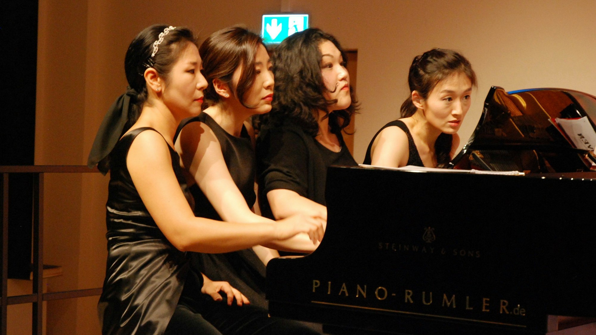 Vier Pianistinnen spielen, nebeneinander sitzend, auf einem Konzertflügel, den Blick konzentriert auf die Noten gerichtet.